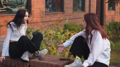 Two girlfriends are arguing, sitting on a street bench apart, young women are in Stock Footage 230984419