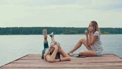 Two girlfriends are lying on the pier near the riverbank and are calling Stock Footage 115649634