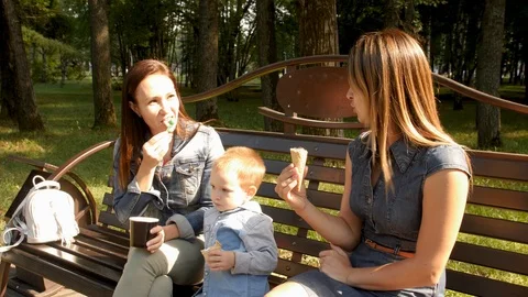 Two girlfriends with a child sit on a bench in the city Park and eat ice cream Stock Footage 94051465