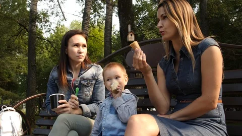 Two girlfriends with a child sit on a bench in the city Park and eat ice cream Stock Footage 94051741
