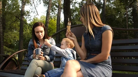 Two girlfriends with a child sit on a bench in the city Park and eat ice cream Stock Footage 94051835