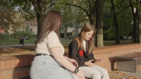 Two girlfriends of different body sizes sitting on a park bench and talking. Vídeos de archivo 307920217