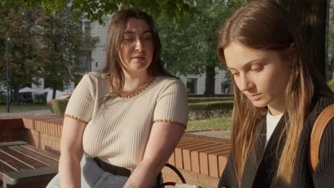 Two girlfriends of different body sizes sitting on a park bench and talking. Stock Footage 307920231
