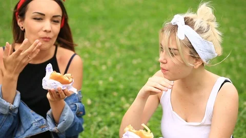 Two girlfriends eat hamburgers, french fries on a green lawn in the city garden Stock Footage
