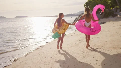 Two girlfriends walk along the beach at sunset holding hands after swimming in Stock Footage 245449452