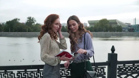 Two girlfriends walk on the bridge flipping through a magazine. 4K Stock Footage 153298853