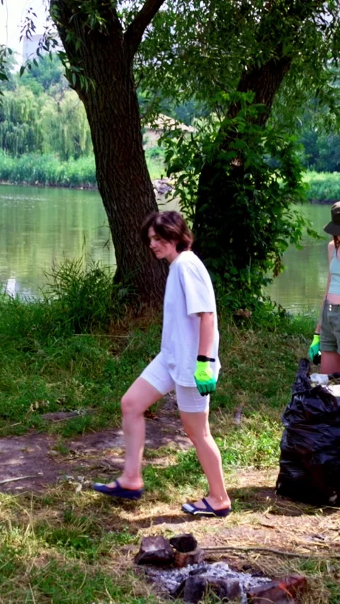 Two girls collecting garbage under big tree near riverbank Stock Footage 340576904