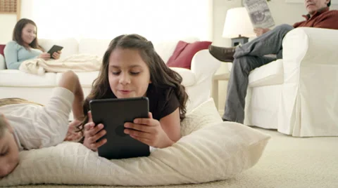 Two girls lie on the floor playing with a tablet as there parents relax Stock Footage 34014164