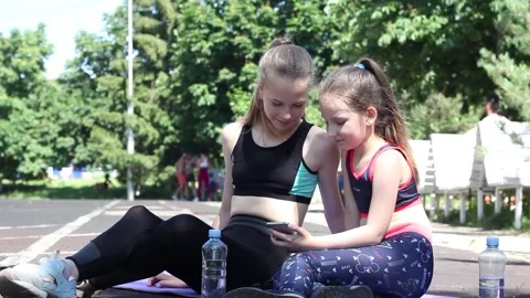 Two girls look at a smartphone while sitting on a mat on a sports ground Stock Footage 132894545
