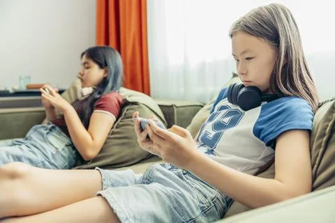 Two girls relaxing on sofa using smartphone, headphones, casual home setting Stock Photos