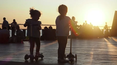 Two girls riding scooter while people walking along the pier and enjoying sunset Stock Footage 128545993