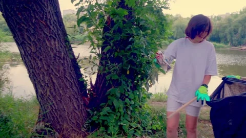 Two girls standing under large tree on riverbank with trash bag Stock Footage 340464121