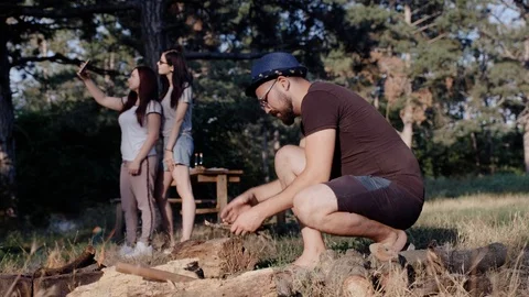 Two girls take a selfie while the guy in the foreground is engaged in a campfire Stock Footage 115043310