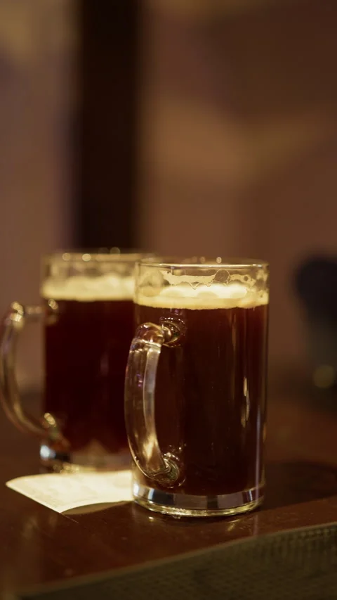 Two glass mugs of dark beer with creamy foam on warm bar table in soft ambient Stock Footage 345266487