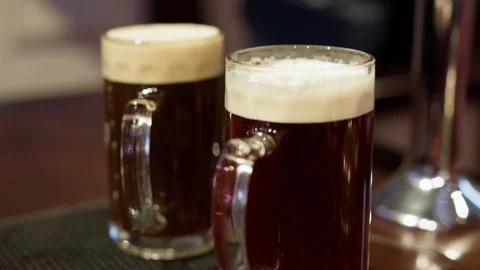 Two glasses of dark beer with creamy foam on bar counter in warm pub atmosphere Stock Footage 345270724