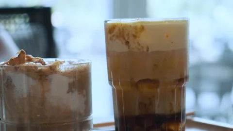 Two glasses of iced creamy coffee drinks with foam and toppings on wooden tray Stock Footage 312813947