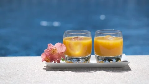 Two glasses with tropical, mango, orange juice with ice cubes on swimming pool Stock-Footage 243995376