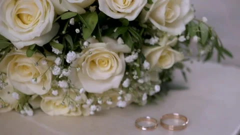 Two gold wedding bands on marble surface next to elegant bridal bouquet Stock Footage 330477561