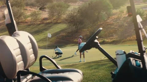 Two golfers reading a putt on a desert green seen past a parked golf cart at Stock Footage 340130392