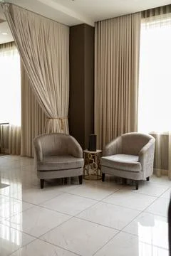 Two gray chairs sit by a small table in a well lit room with large windows .. Photos