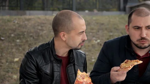 Two guys are sitting in the park and eating seafood pizza Stock Footage 97799688