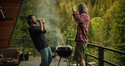 Two guys dance and have fun while cooking on the grill on a spacious balcony Stock Footage 246416326