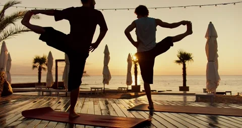 Two guys do yoga and stand on one leg on the beach in the morning. Silhouettes 動画素材 249435479
