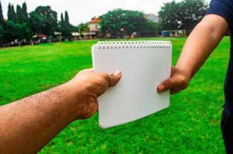 Two hands are seen exchanging a blank spiral notebook in an outdoor park se.. Stock Photos