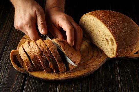 Two hands cut a loaf of bread into slices on a wooden board. The scene takes  Stock Photos