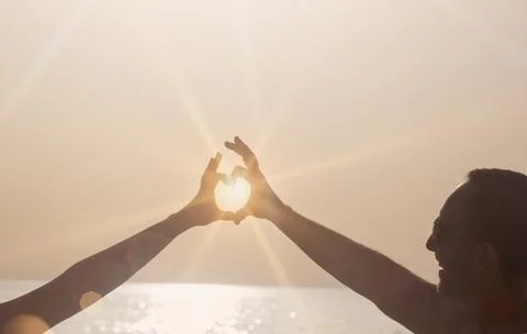 Two hands form a heart shape against a bright sun with rays above a calm sea  Fotos Stock