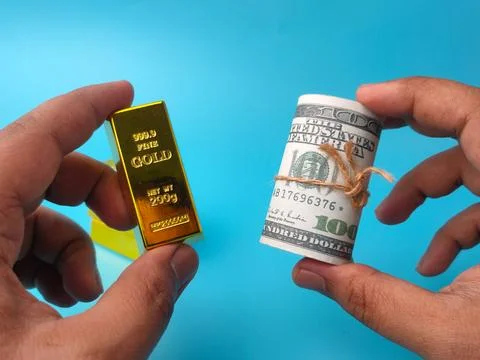 Two hands frame a shiny gold bullion bar on the left and a rolled stack of .. Stock Photos