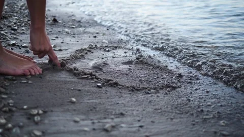 Two hands gracefully draw a heart on the sand by the waters edge each stroke Stock Footage 282698475