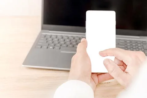 Two hands grip a white-screen phone above a dark laptop on a light wood desk. Stock Photos