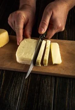 Two hands hold a knife and slice a block of cheese on a wooden cutting boar.. Stock Photos