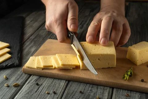 Two hands hold a knife to cut a block of cheese on a wooden cutting board. .. Stock Photos