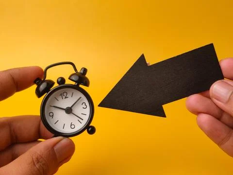 Two hands hold a small black alarm clock and a black paper arrow pointing a.. Stock Photos