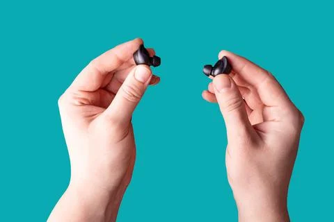 Two hands holding black wireless earbuds against a teal background Stock Photos