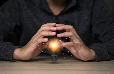 Two hands protecting the light bulb that is illuminating on the table. Stock Photos