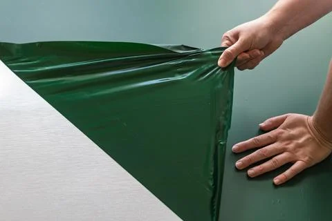Two hands pull a wide sheet of green vinyl from a light kitchen cabinet door Stockfoto's