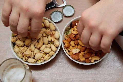 Two hands taking nuts from plates next to a beer glass and a bottle opener, b Stock Photos