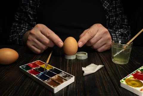 Two hands work on decorating an egg with paint during a craft session. A pain Stock Photos