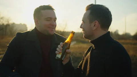 Two handsome men stand in a field with bottles of alcohol, they chat and laugh.  Stock Footage 121613067