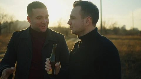 Two handsome men stand in a field with bottles of alcohol, they chat and laugh.  Stock Footage 121613407