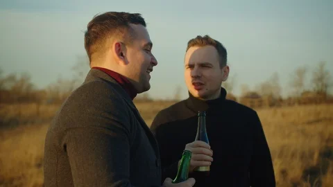 Two handsome men stand in a field with bottles of alcohol, they chat and laugh.  Stock Footage 121613415