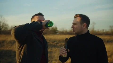Two handsome men stand in a field with bottles of alcohol, they chat and laugh.  Stock Footage 121621596