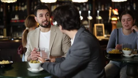 Two handsome positive young guys friends talking and drinking beer at table in Stock Footage 341382686