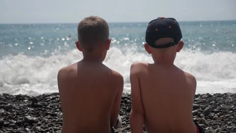 Two happy boys are sitting on the seashore, the waves are slowly rolling in. Stock Footage 333246429