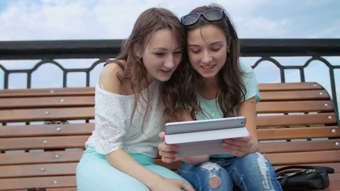 Two happy girs sitting on the bench near the river, using tablet device, looking Stock Footage 74482776