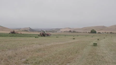 Two hay making tractors drive slowly across farm landscape Stock Footage 332255832