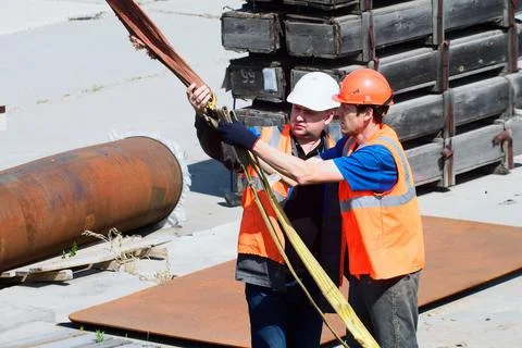 Two helmeted slingers attach load to slings outside on summer afternoon. Loading Stock Photos